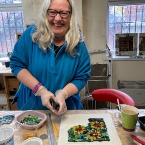 workshop participant smiling whilst using tile nippers to cut material for a mosaic. The mosaic is visible on the table in front of her as a work in progress.