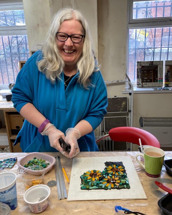 workshop participant smiling whilst using tile nippers to cut material for a mosaic. The mosaic is visible on the table in front of her as a work in progress.